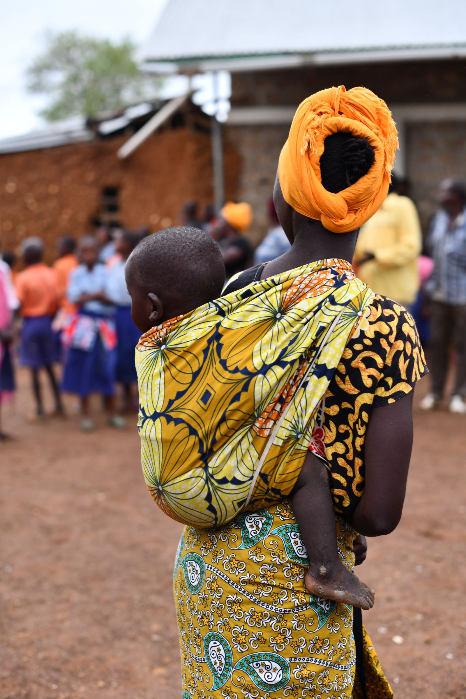 A mother carries her child on her back in a bright yellow patterned wrap.