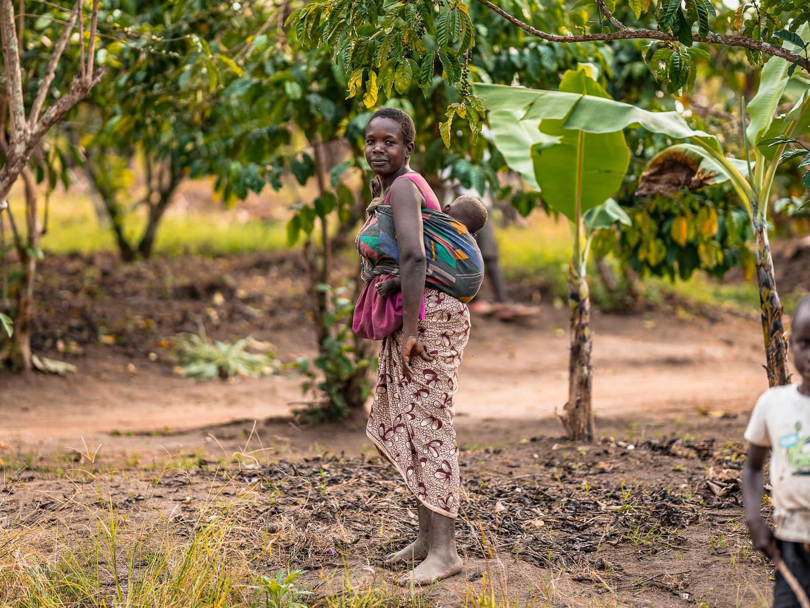 A woman carrying her baby on her back in a green village garden.